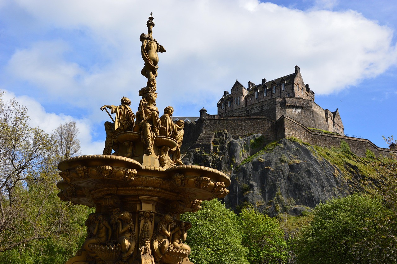 La Evolución del Castillo de Edimburgo_ De fortaleza a icono nacional Golden fountain statues with a castle on a hill in the background, surrounded by trees under a blue sky.
