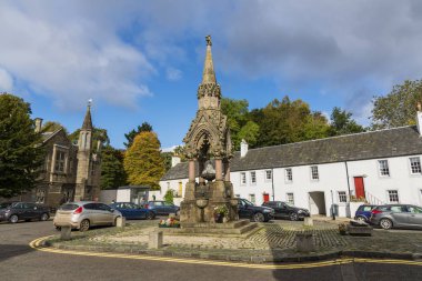 Historic monument with ornate spire in a square, surrounded by cars and white buildings.