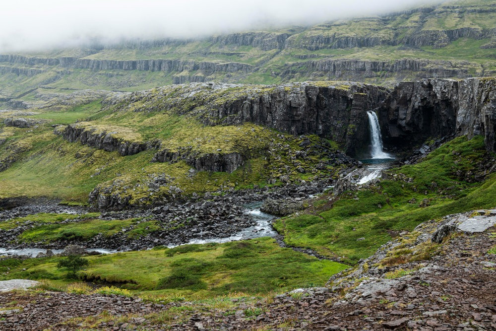 Waterfall cascading in a rocky green landscape shrouded in clouds.