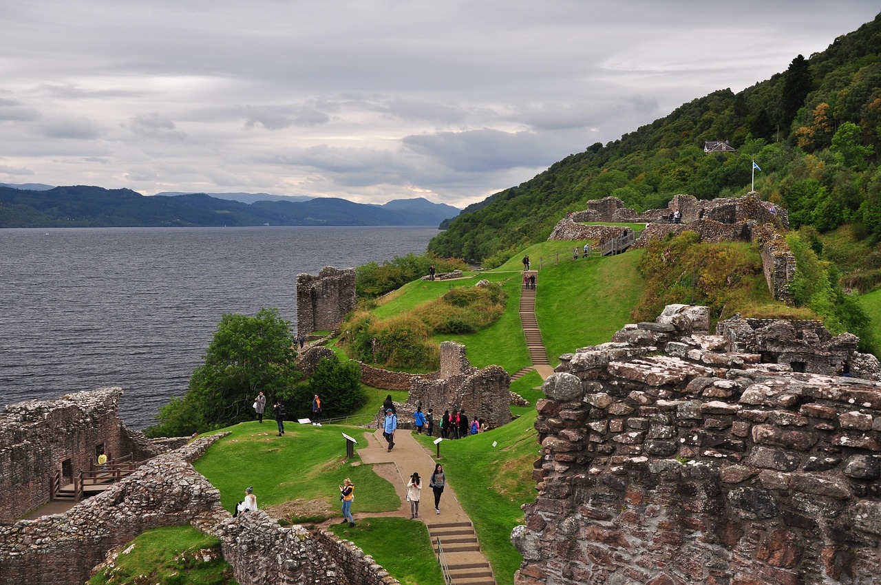 Ruins of a castle by a lake with tourists, surrounded by greenery and hills under a cloudy sky.