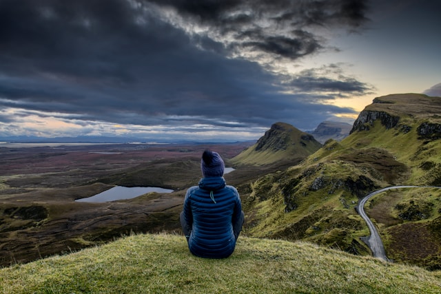 Person in blue jacket sitting on hill overlooking a scenic landscape with mountains and cloudy sky.
