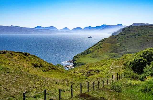 Scenic view of grassy hills, ocean, and distant mountains under a clear blue sky.