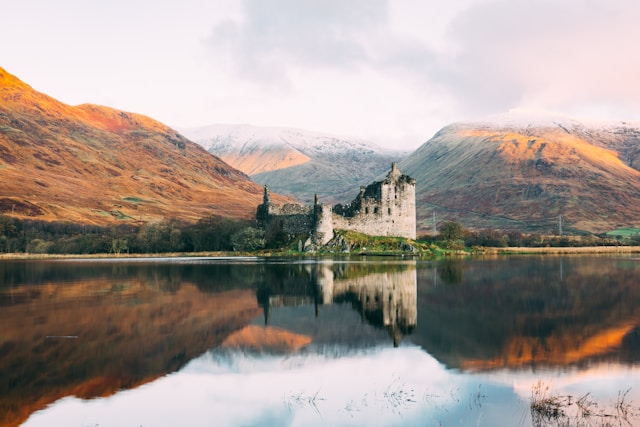 Ruined castle by a lake with mountains in the background at sunset.