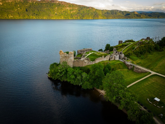 Aerial view of a castle ruin by a large lake with lush green surroundings.