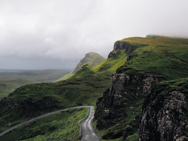 Winding road through lush, green hills with misty clouds overhead.