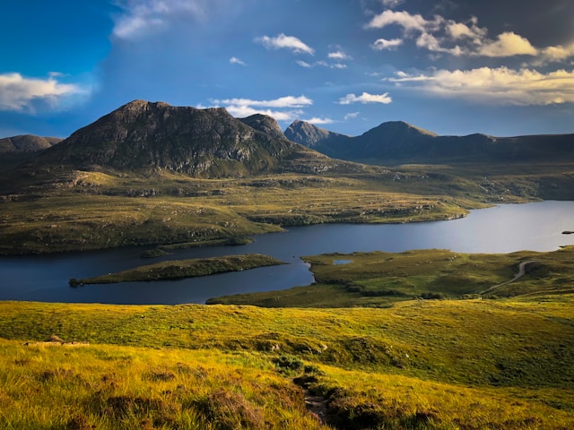 Scenic landscape with a lake, grassy hills, and distant mountains under a blue sky.
