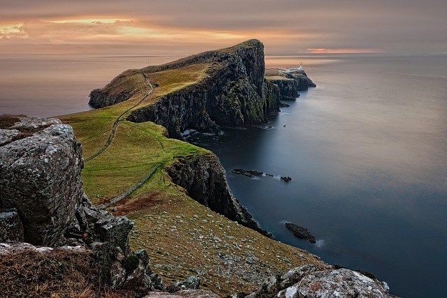 Rocky coastline with grassy cliffs and a lighthouse at sunset.