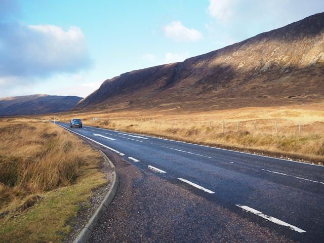Car on a rural road with mountains and grasslands under a partly cloudy sky.